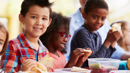 School-age children eating sandwiches together and one child is smiling at the camera.