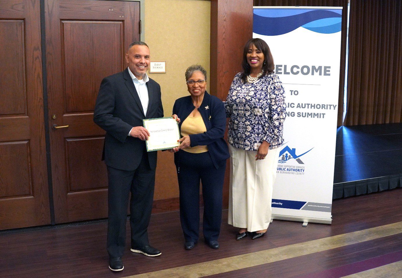 A play button icon for a video placed over a photo of a man in a dark suit and a woman in white pants and a purple and white top standing in front of an IHSS Public Authority Caregiver Training Summit banner, presenting a certificate to a woman in a navy suit.