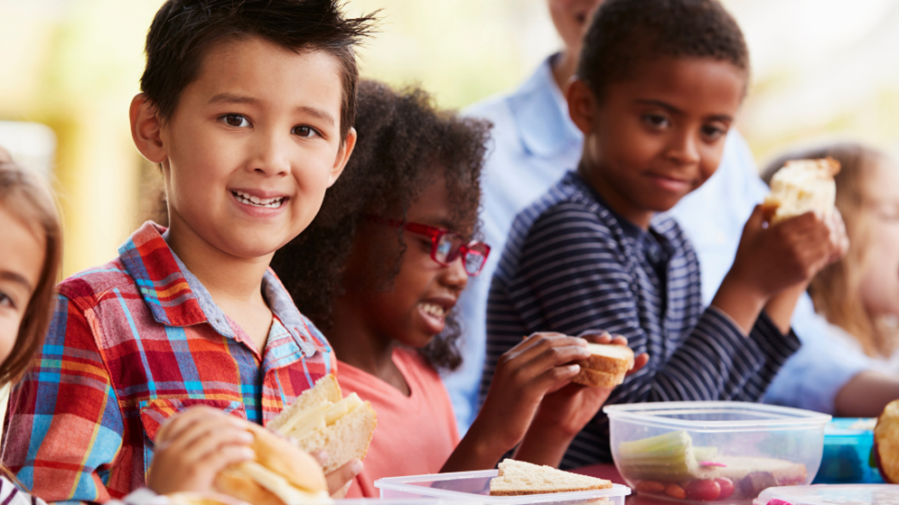 School-age children eating sandwiches together and one child is smiling at the camera.