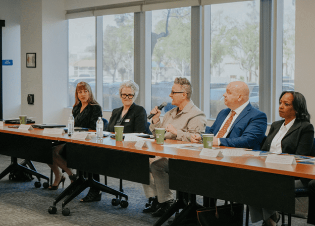 A group of five people sit at a conference table in a well-lit room with large windows. One person holds a microphone, suggesting a discussion.