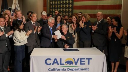 Governor Newsom is seated at a table with CALVET California Department of Veterans Affairs tablecloth, holding a signed bill with a group of people applauding behind him.
