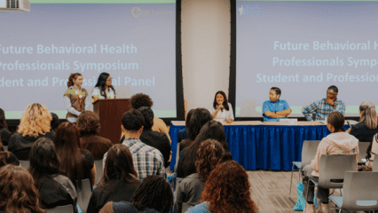 A panel of five speakers addresses an audience at the Future Behavioral Health Professionals Symposium, with event details projected on screens behind them.