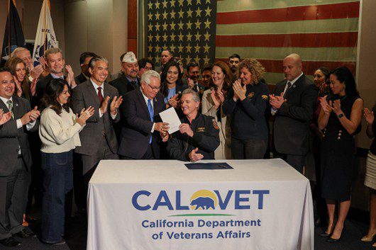 Governor Newsom is seated at a table with CALVET California Department of Veterans Affairs tablecloth, holding a signed bill with a group of people applauding behind him.