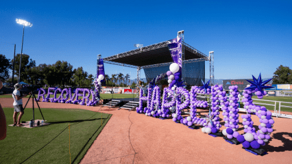 An outdoor stage at a recovery event, decorated with purple and white balloons spelling out "Recovery Happens."