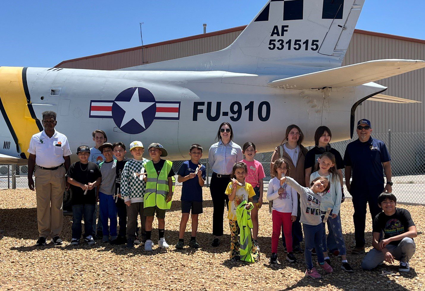 A group of children and adults pose for a photo in front of a historic U.S. Air Force jet with the tail number "AF 531515" and body number "FU-910." The aircraft is parked outdoors near a hangar, and the group stands smiling under bright sunlight.
