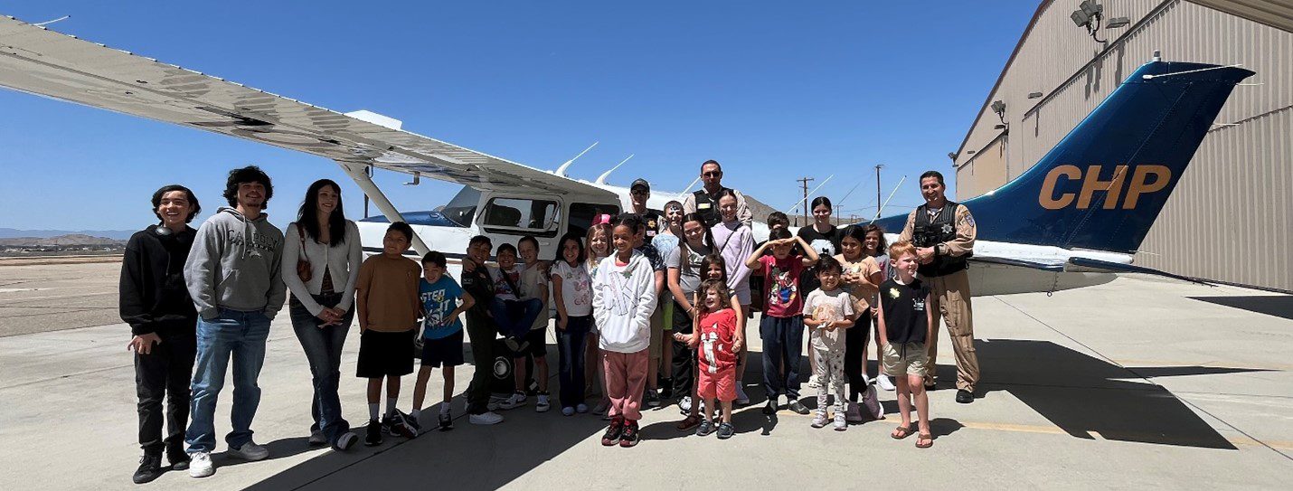 A large group of children and adults pose for a group photo in front of a California Highway Patrol airplane parked on the tarmac beside a hangar. The group smiles under a clear blue sky, with the aircraft’s tail marked "CHP" prominently visible.