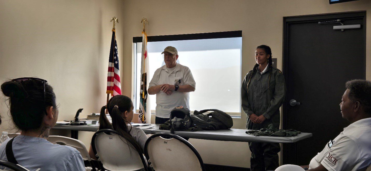 A small classroom setting where a uniformed instructor and a person in a fighter pilot suit stand at the front, presenting flight gear laid out on a table to an attentive seated audience. The room includes American and Californian flags, and some of the gear includes a helmet and flight suit accessories.