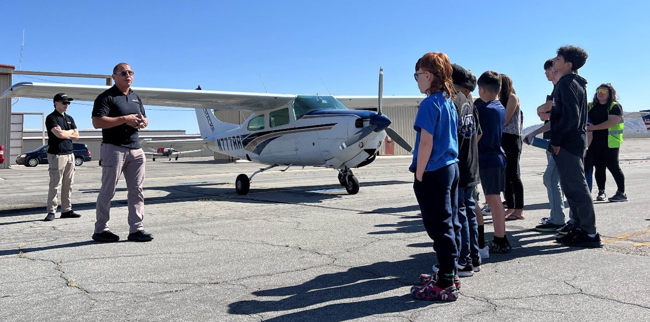 A group of children stands in a line on an airport tarmac, listening to a man in sunglasses speak near a small white airplane. Another adult stands in the background, and the scene takes place under a clear blue sky with hangars visible in the distance.