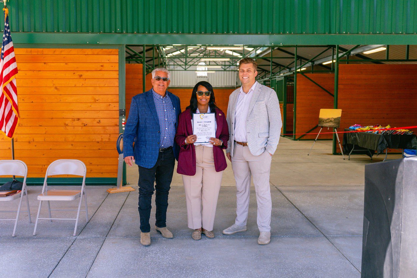 Two men flank a woman while she holds a certificate in front of open barn doors with the American flag and some metal chairs in the background.