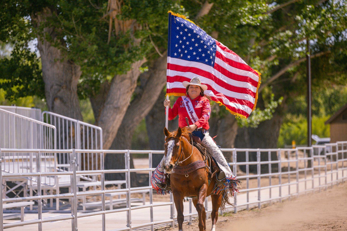A woman dressed in Western attire carries the American flag while riding a horse in a dirt arena.