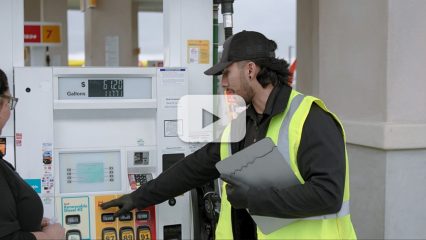 A man dressed in black uniform with ball cap and fluorescent yellow vest holds a clip board and points to a fuel pump dispenser at a gas station while a woman wearing glasses looks where he points.