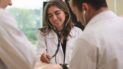 Smiling female doctor in a white coat using a stethoscope to examine a patient’s arm while two other medical professionals stand nearby during a clinical checkup.