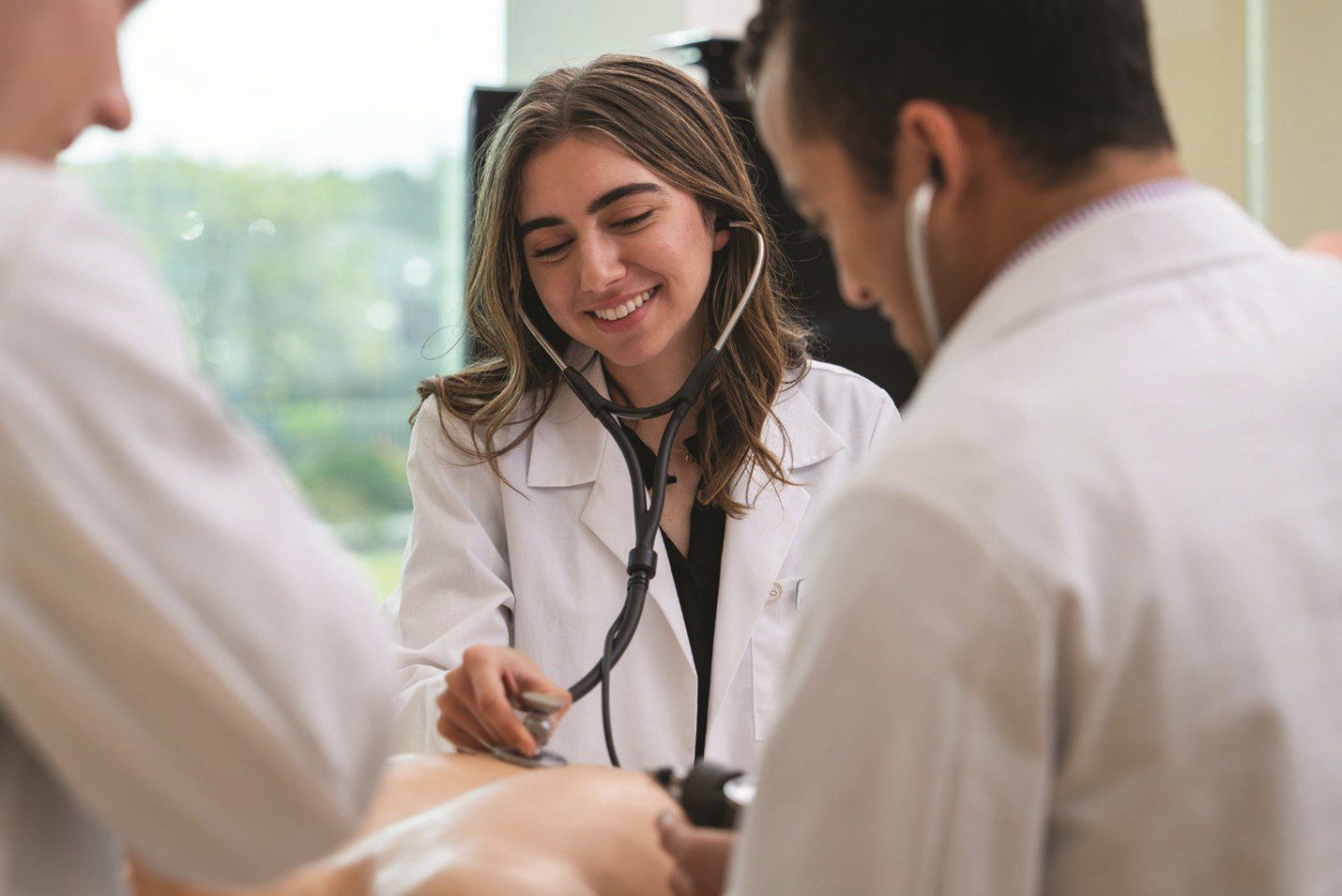Smiling female doctor in a white coat using a stethoscope to examine a patient’s arm while two other medical professionals stand nearby during a clinical checkup.