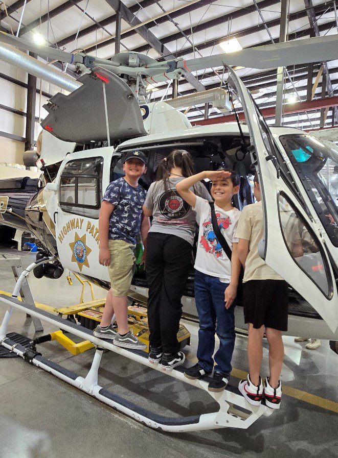 Students stand on the skid of a California Highway Patrol H-82 helicopter inside a hangar, with some entering the cockpit and one child playfully saluting the camera.