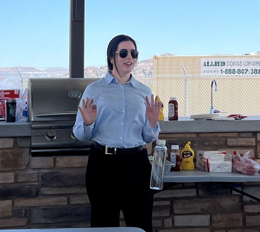 A woman in business casual clothes and sunglasses speaks in front of an outdoor grill.