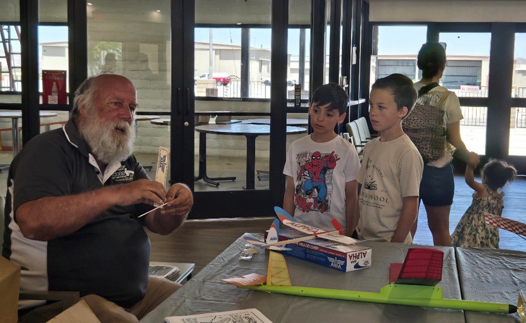 Ronnie McCullough sits at a table indoors, demonstrating how to assemble a model airplane to two young boys watching attentively. Several model airplane kits and parts are spread across the table. In the background, other children and adults move through the space.