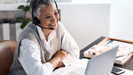 Person speaking with a woman on a computer, white cell phone top of a wooden desk, blurred background.