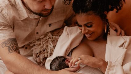 Parent breastfeeds a newborn while another parent sits close beside them.
