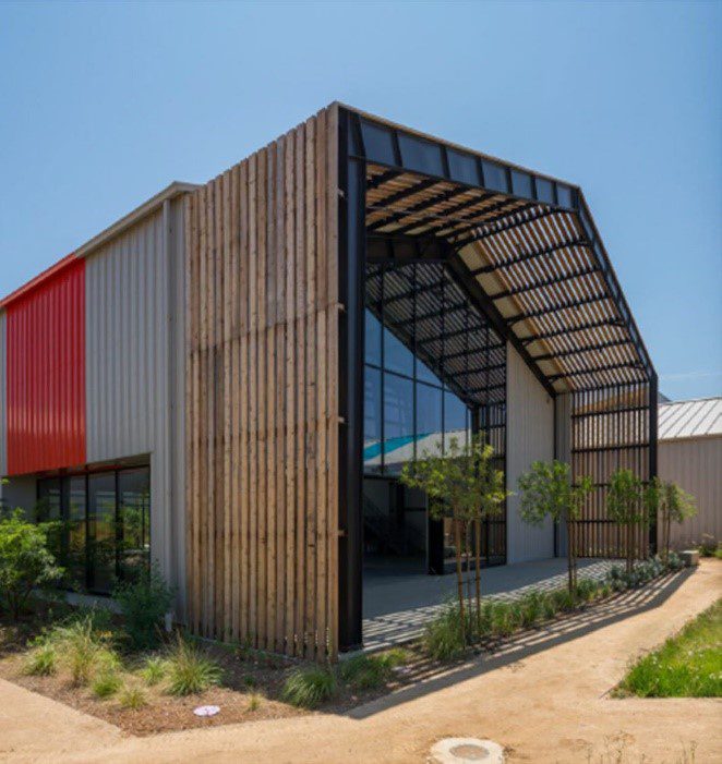 building with metal, wood framing and glass windows are surrounded by desert landscaping with a dirt pathway.