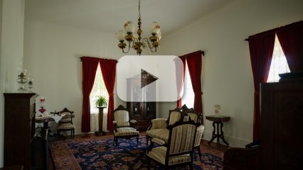A vintage-style living room with furniture, a chandelier, a cabinet and an area rug. A video play button icon overlays the image.
