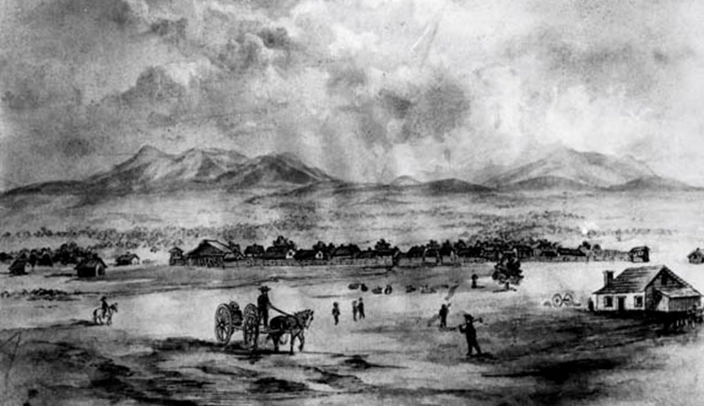 Black and white photo featuring a small town with houses, settlers and a horse-drawn cart with mountains in the background.