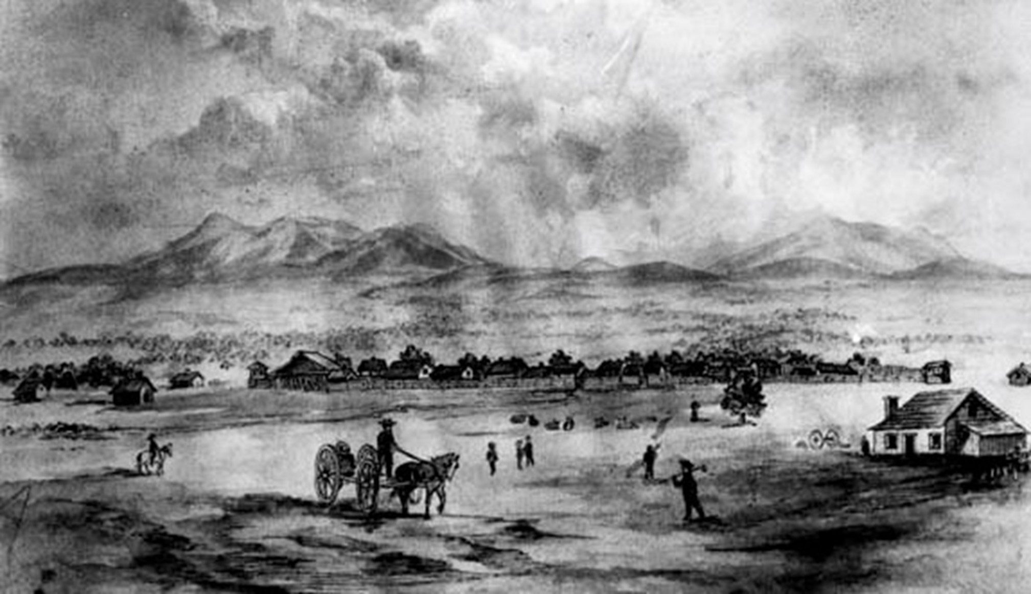 Black and white photo featuring a small town with houses, settlers and a horse-drawn cart with mountains in the background.