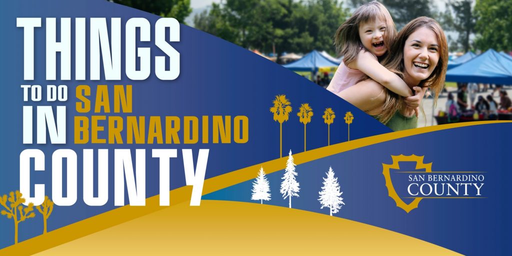 A banner reading 'Things to Do in San Bernardino County' featuring a vibrant design with blue and gold accents. On the right, a woman and a laughing child enjoy a sunny day at an outdoor event with tents in the background. The San Bernardino County logo is included, along with stylized tree silhouettes.