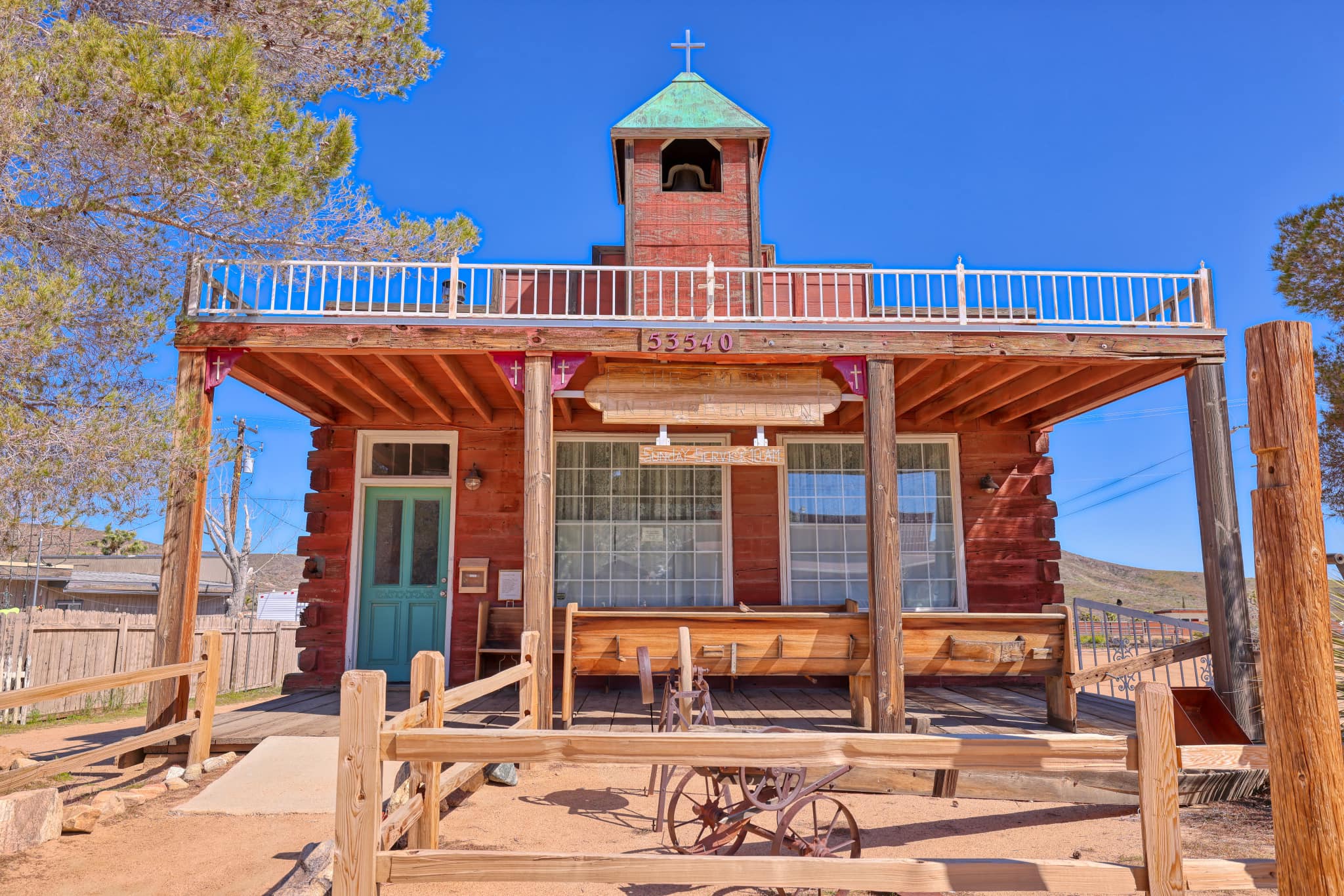 Exterior view of a church made of red wood surrounded by a wooden fence in a desert landscape.
