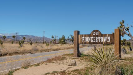 A wooden sign reading “1946 Pioneertown” stands beside a rural desert road with dry vegetation, Joshua trees and distant mountains under a clear blue sky.