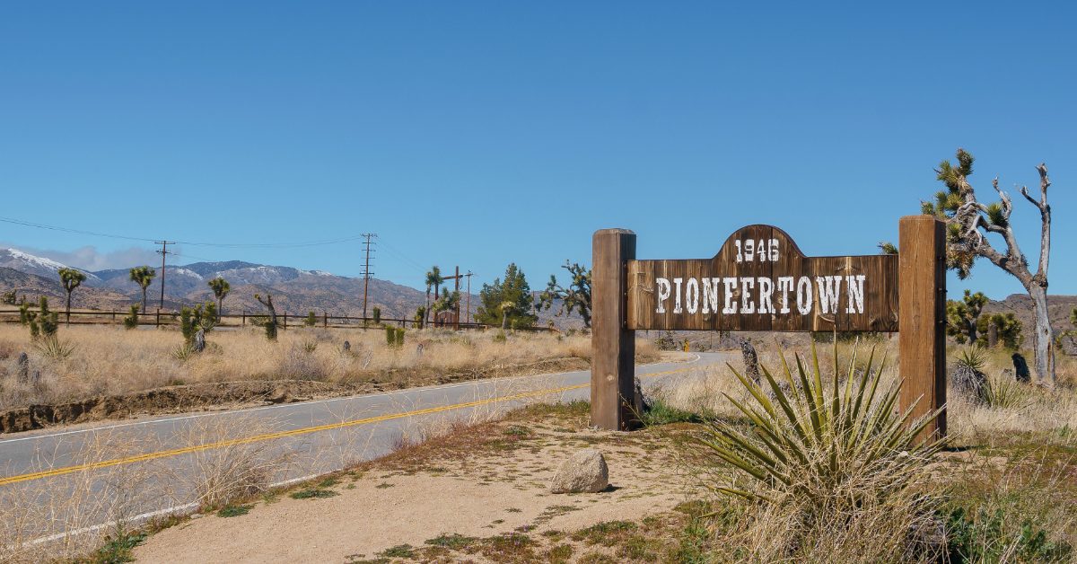 A wooden sign reading “1946 Pioneertown” stands beside a rural desert road with dry vegetation, Joshua trees and distant mountains under a clear blue sky.
