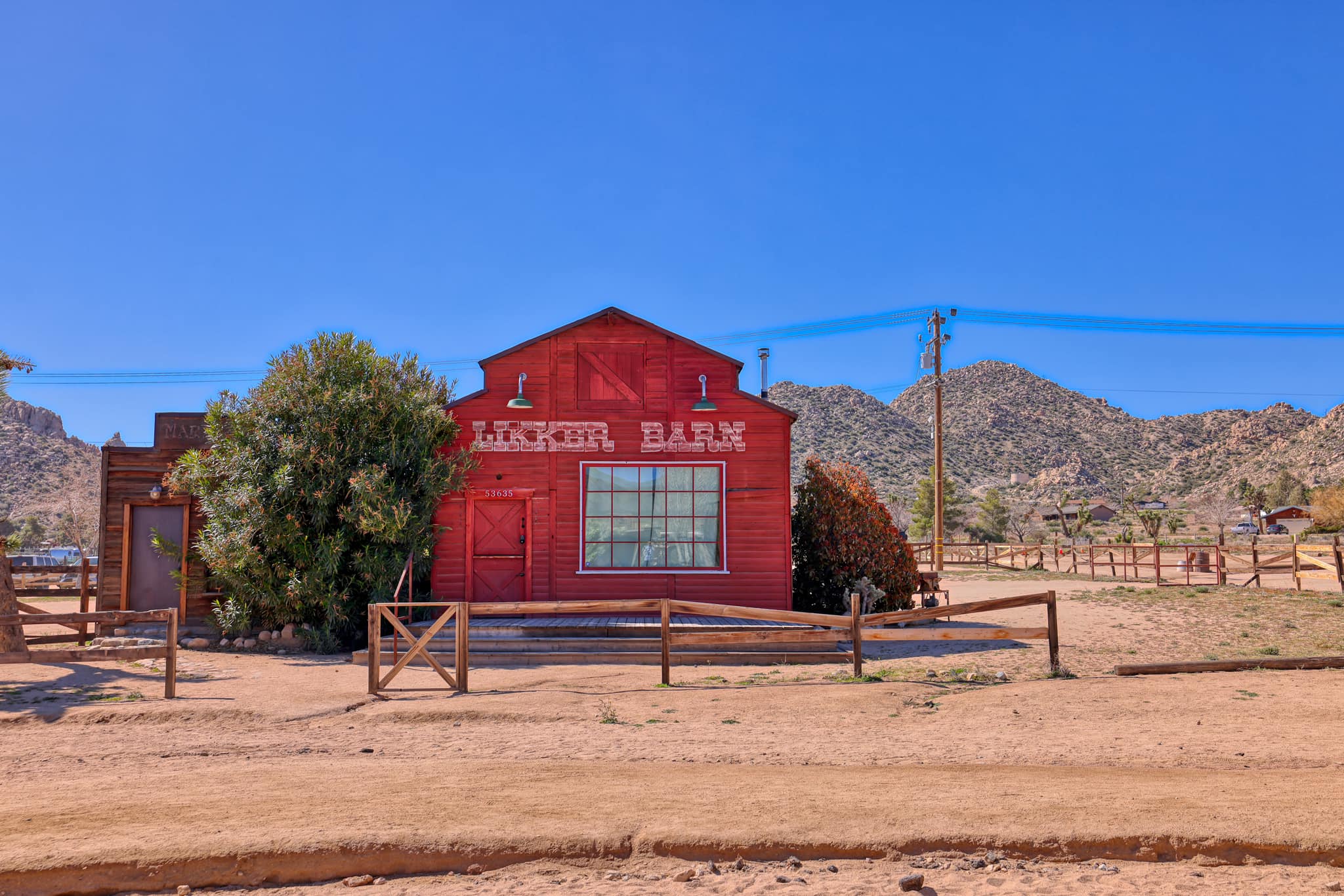 View of the Pioneertown General Store, which is a red wooden building with a large front window set in a desert landscape with very little vegetation and distant hills.