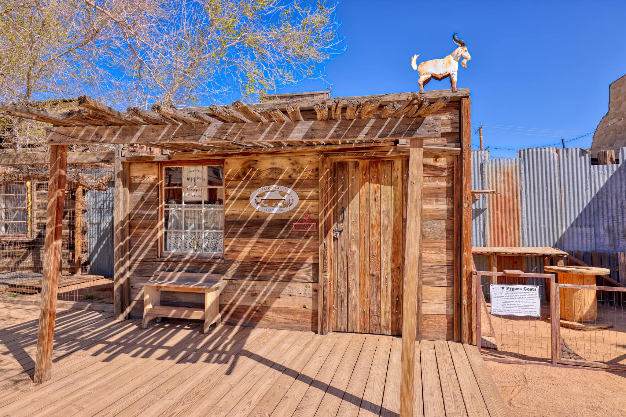 Exterior view of a rustic wooden building featuring a goat statue on the roof and surrounded by a wooden fence and benches.