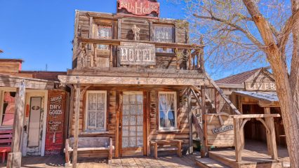 Image of a wooden building with signs reading "Bath House" and "Hotel." Next door is another building with a sign that says, "Dry Goods.” The structures have a vintage, Old West architectural style.