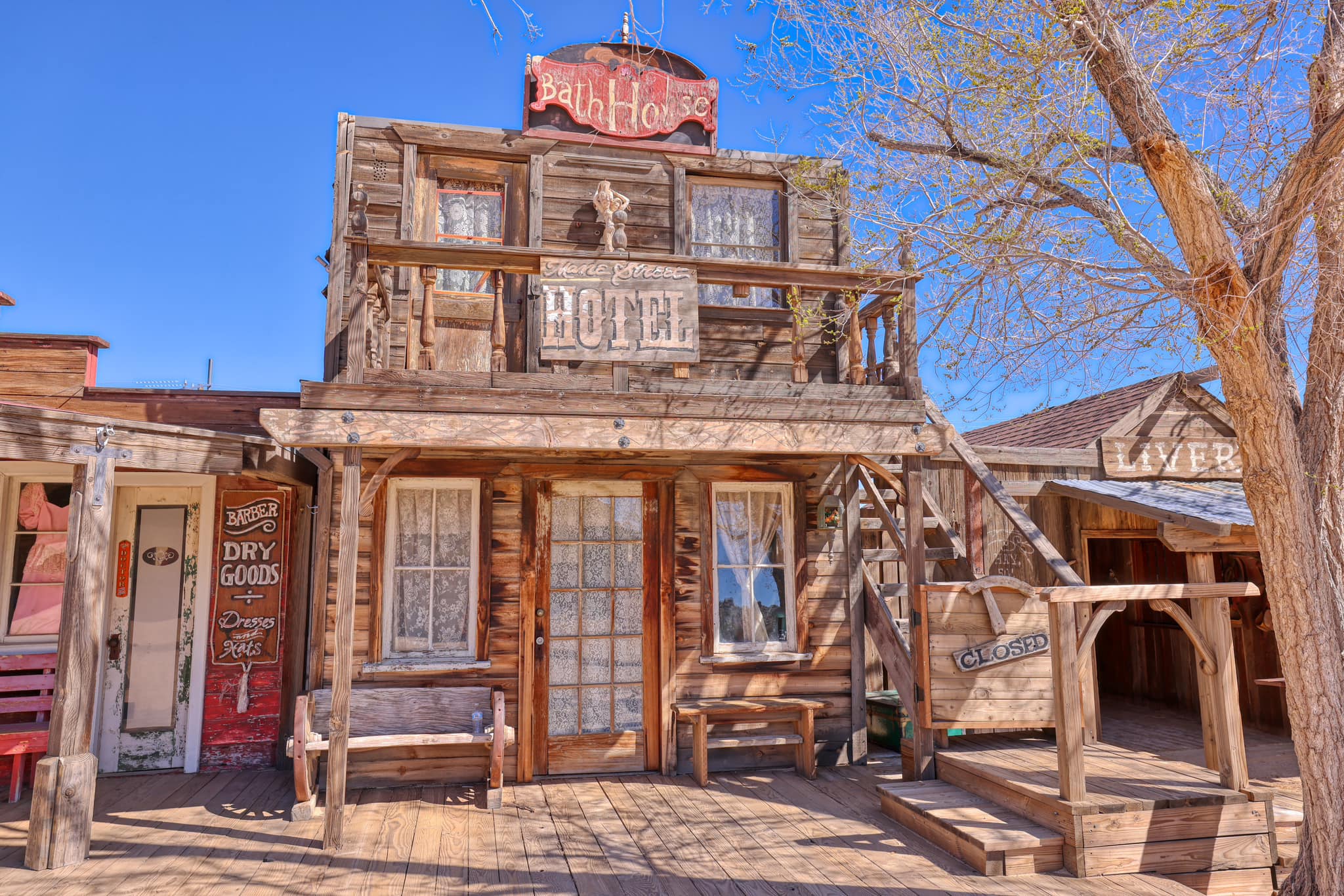 Image of a wooden building with signs reading "Bath House" and "Hotel." Next door is another building with a sign that says, "Dry Goods.” The structures have a vintage, Old West architectural style.