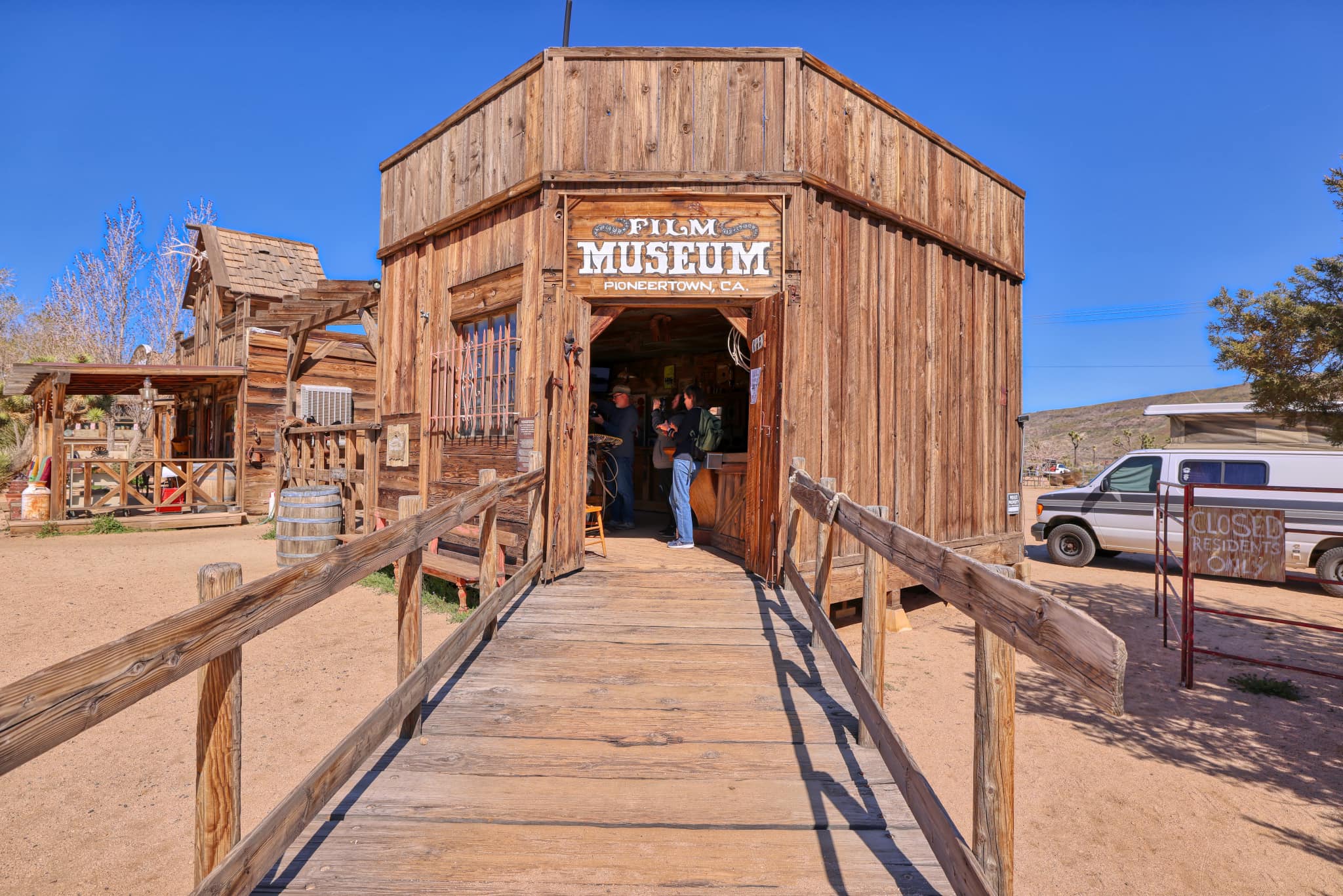 Exterior view of the Film Museum in Pioneertown. It is a rustic wooden building with a ramp leading up to the entrance. Several individuals are visible near the entrance.