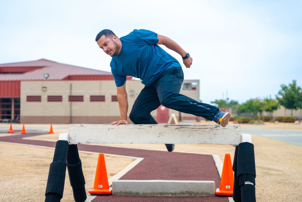 Man in blue shirt and black pants climbing over a wooden obstacle on an outdoor course.