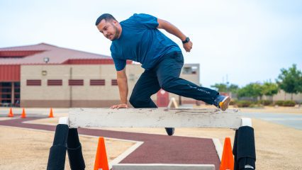 Man in blue shirt and black pants climbing over a wooden obstacle on an outdoor course.