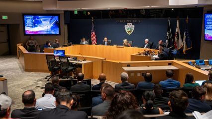 The Board of Supervisors meeting in progress with members seated at the dais under the motto "In God We Trust" and surrounded by flags with an audience in the background.