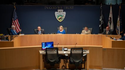Several individuals seated at a dais. Behind them is a navy-blue wall with the phrase "In God We Trust" and the county seal.