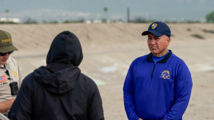 Two individuals engage in conversation with a homeless person who is standing by a tent in an encampment area.
