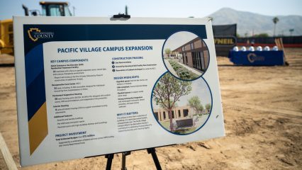A sign labeled "Pacific Village Campus Expansion" at a construction site that includes campus components, a project investment summary, phasing, design highlights and why it matters.