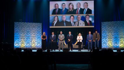 A group of seven individuals standing on a stage at a public event, with a background screen displaying additional images of these people and others.