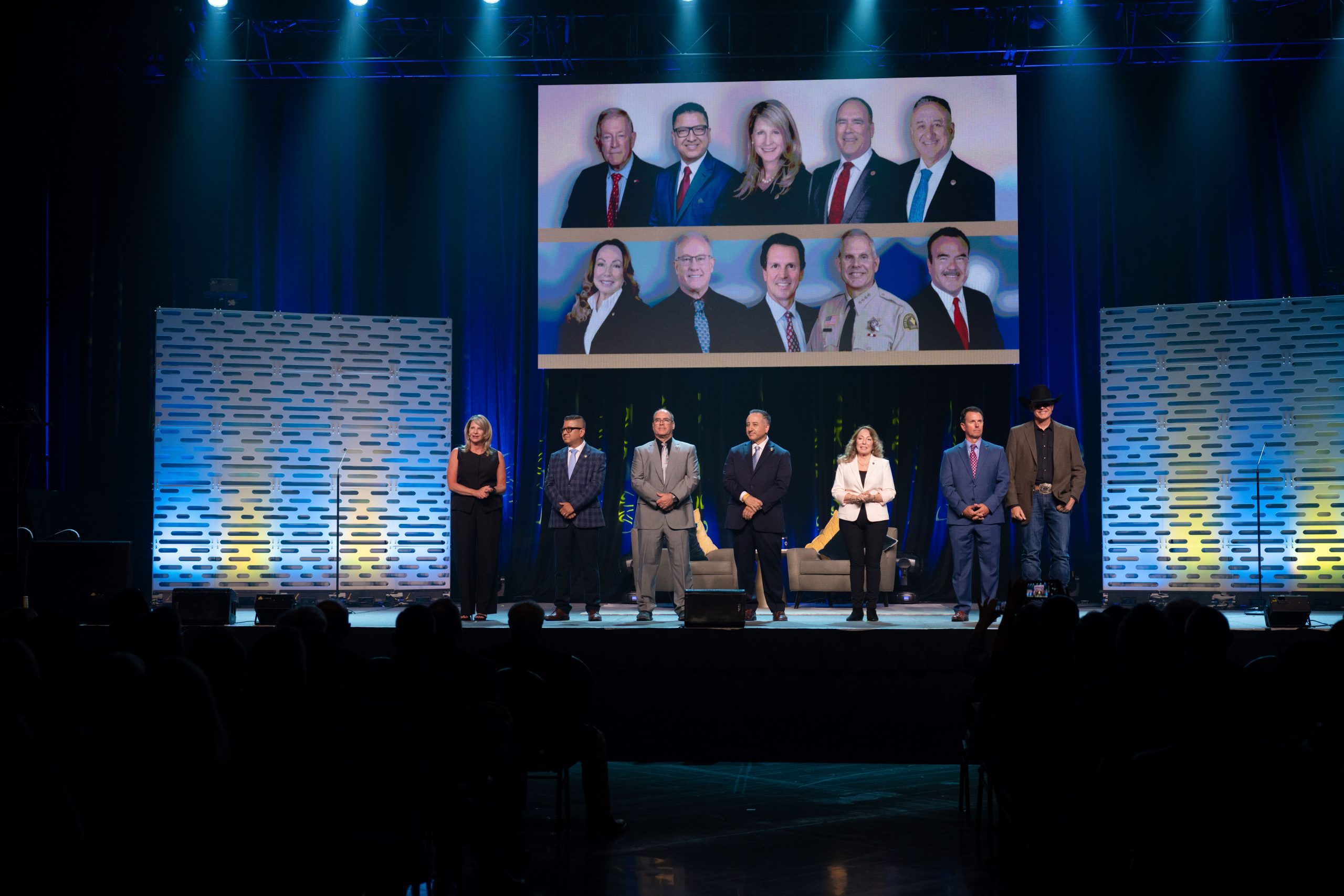 A group of seven individuals standing on a stage at a public event, with a background screen displaying additional images of these people and others.