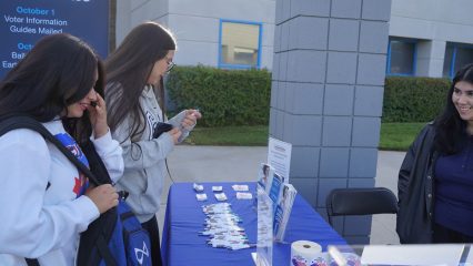 Two students at Rialto High School smile and talk with a Registrar of Voters staff member in front of a table with blue linen cloth filled with stickers, pens, mints and flyers.