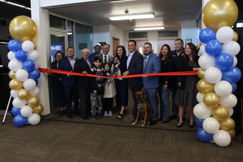 A ribbon-cutting ceremony with a group of smiling attendees, including Vice Chairman and Fifth District Supervisor Joe Baca, Jr., members of the Rialto City Council, community leaders, children and a service dog. The event takes place in front of a decorated doorway in the Rialto Library with gold, white and blue balloons, as the County Librarian Melanie Orosco cuts the red ribbon with large scissors.