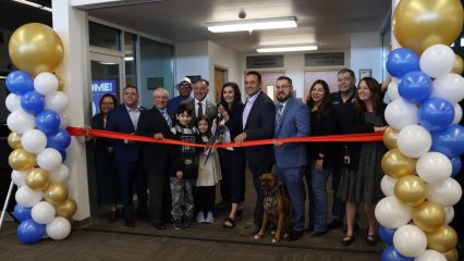 A ribbon-cutting ceremony with a group of smiling attendees, including Vice Chairman and Fifth District Supervisor Joe Baca, Jr., members of the Rialto City Council, community leaders, children and a service dog. The event takes place in front of a decorated doorway in the Rialto Library with gold, white and blue balloons, as the County Librarian Melanie Orosco cuts the red ribbon with large scissors.