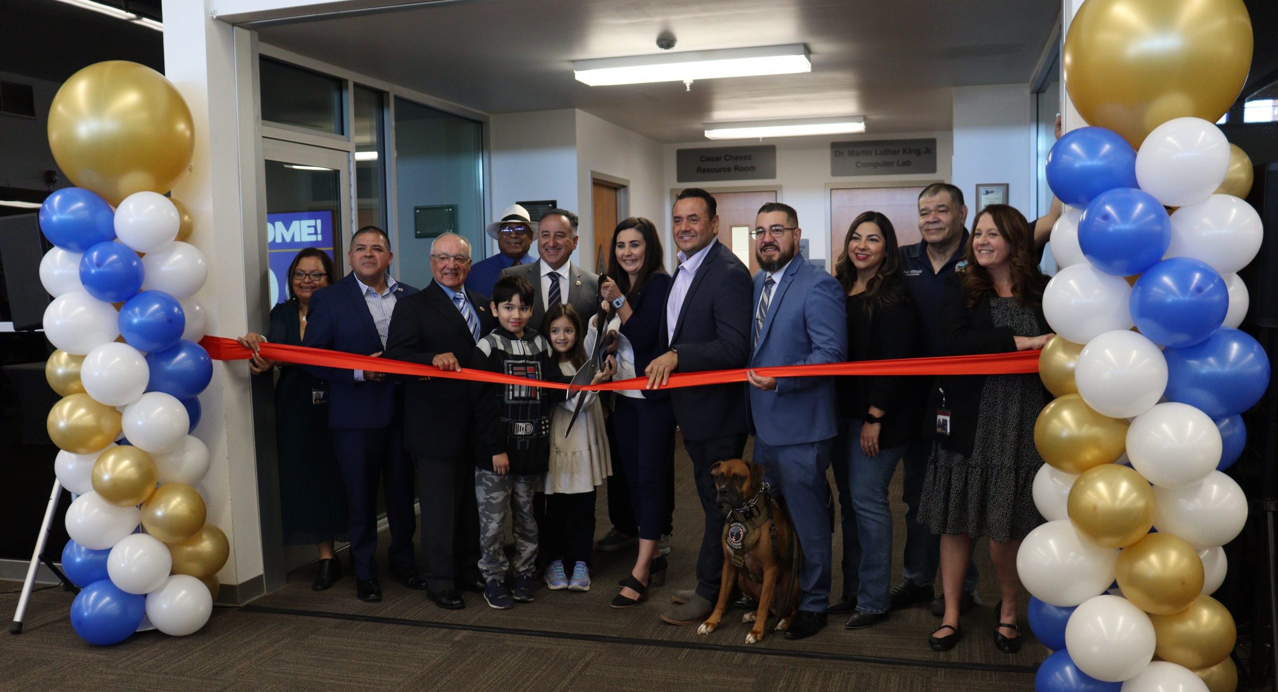 A ribbon-cutting ceremony with a group of smiling attendees, including Vice Chairman and Fifth District Supervisor Joe Baca, Jr., members of the Rialto City Council, community leaders, children and a service dog. The event takes place in front of a decorated doorway in the Rialto Library with gold, white and blue balloons, as the County Librarian Melanie Orosco cuts the red ribbon with large scissors.