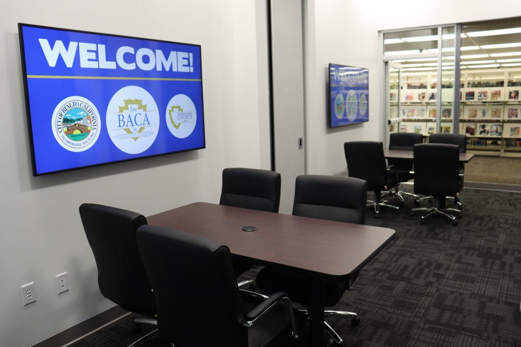 A quiet study room at the Rialto Library, furnished with a dark wood table and black rolling chairs. A large screen on the wall displays 'Welcome!' with county and local city logos, while a glass window provides a view of bookshelves in the main library area.