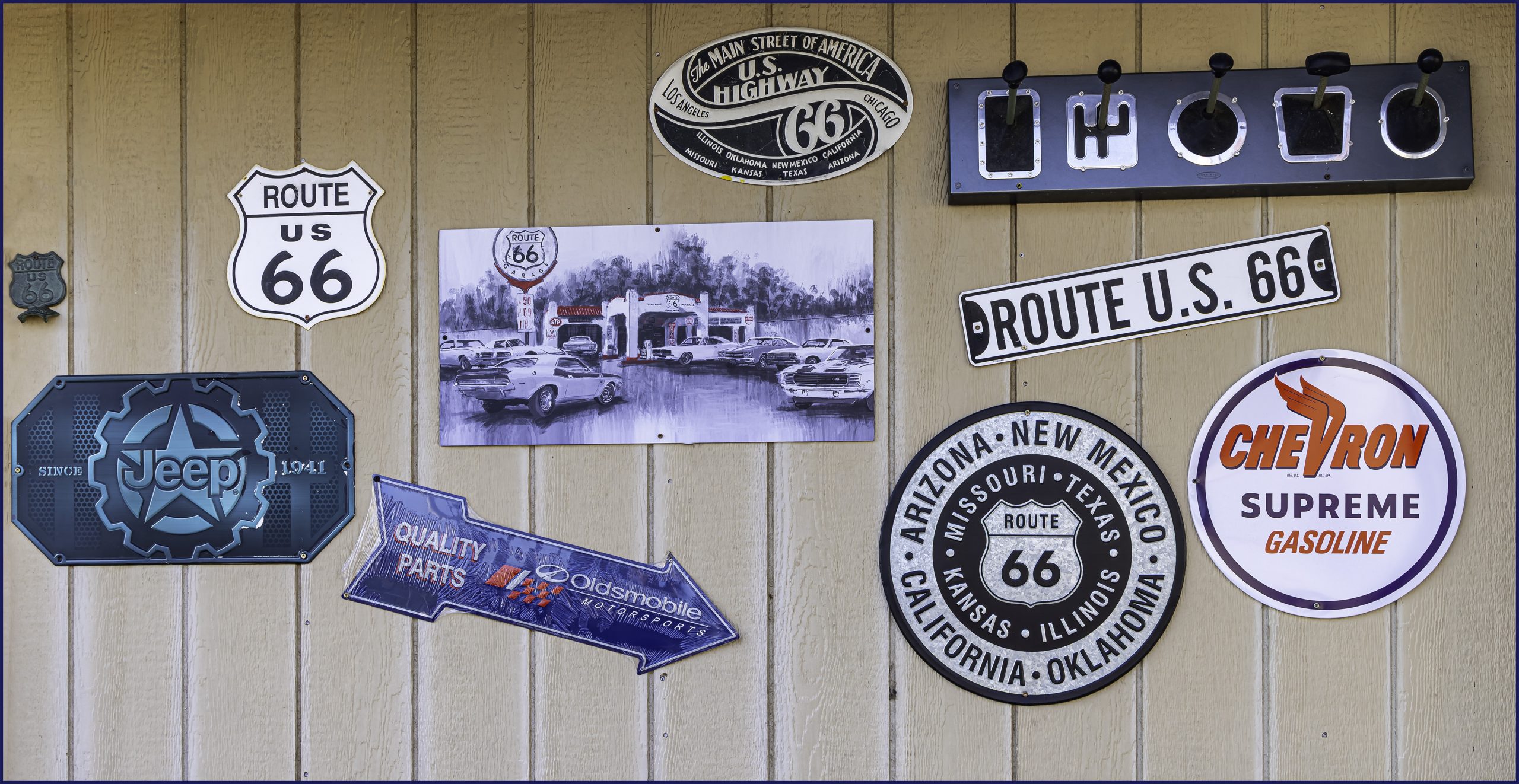 A collection of vintage signs displayed on a wall. The signs include Route 66, Jeep, Chevron Supreme Gasoline, and a photo featuring cars at a retro gas station.