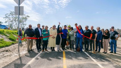 a group of about 20 people cheer and smile while standing on a portion of the Santa Ana River Trail bike path behind a large red ribbon that is cut into two pieces.